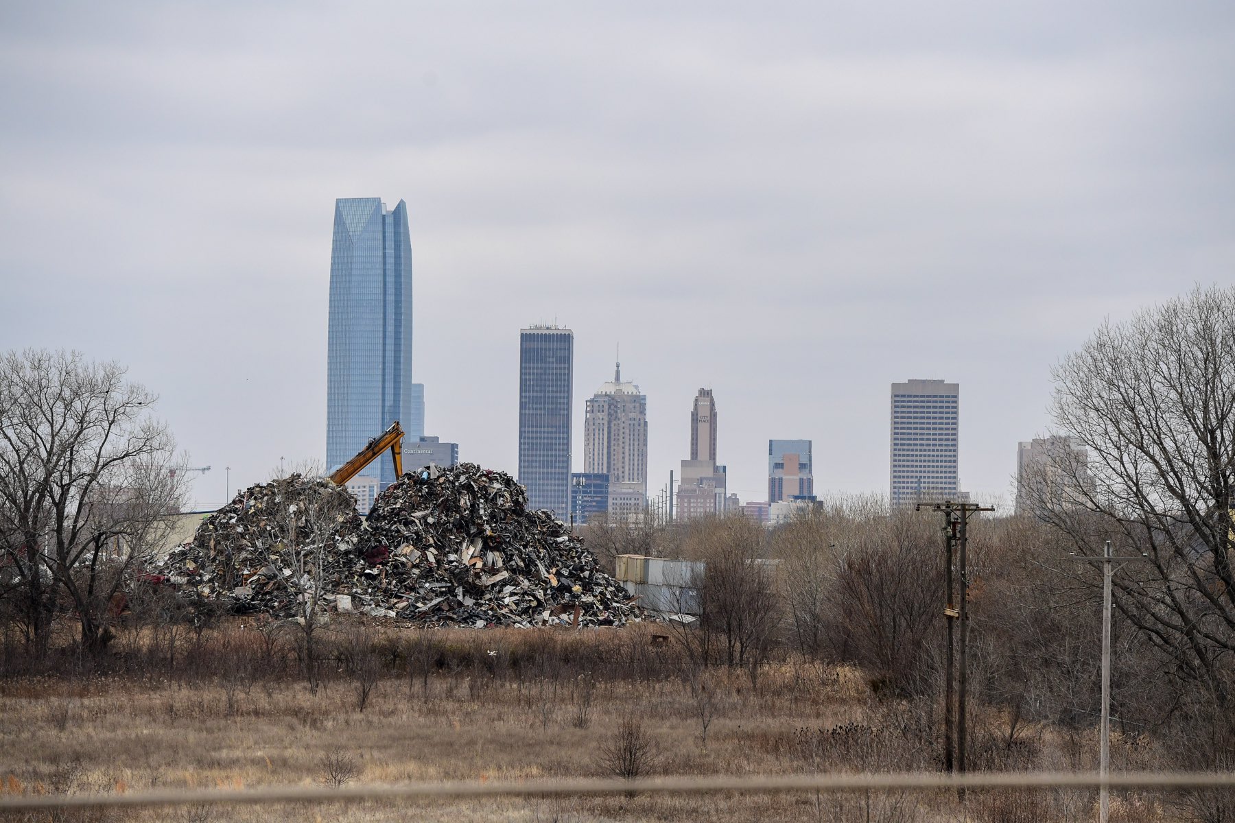 A mountain of crushed scrap metal and an excavator in a field, with the downtown Oklahoma City skyline behind it